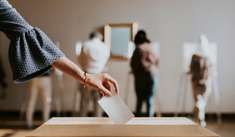 hand-depositing-ballot-with-us-flag-in-background-2026-03-18-13-17-46-utc.png